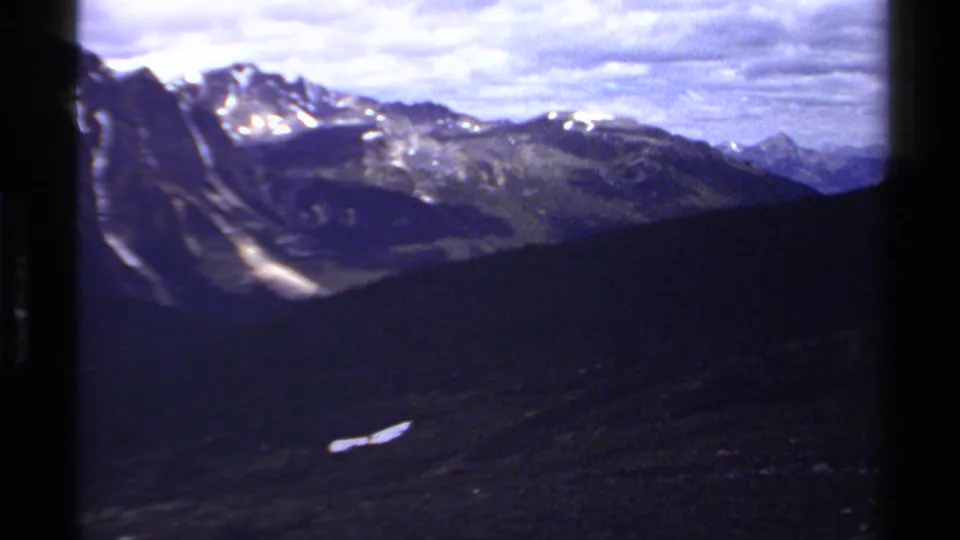 1979 British Columbia mountain vista on 8mm film, with rugged peaks, snow patches, forested slopes, and cloudy sky.