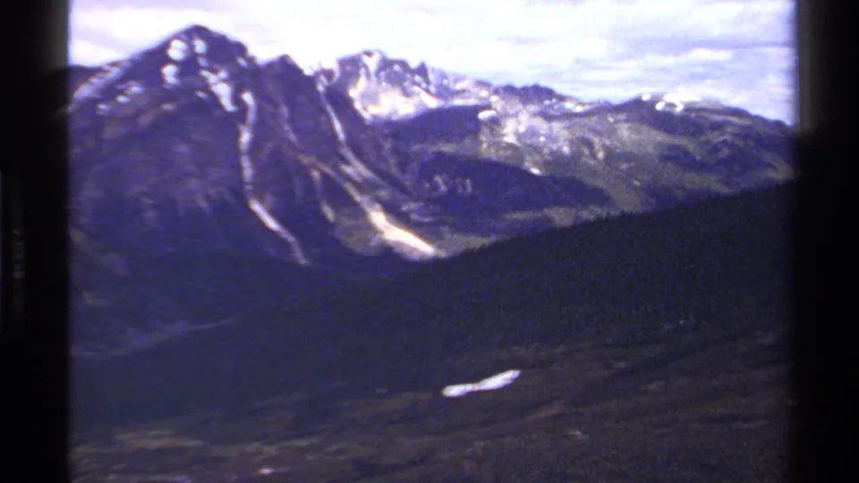 Vintage 8mm view of British Columbia mountains, showing snow-streaked peaks, dark wooded slopes, and a cloudy sky.