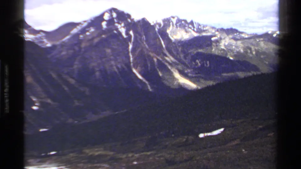 Soft 8mm frame of British Columbia peaks, with lingering snowfields, a cloudy sky, and a dark wooded foreground.