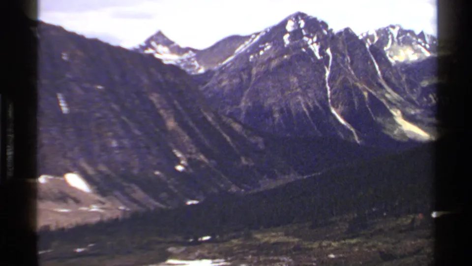 1979 British Columbia mountain view with a central rocky peak, narrow snow streaks, and dark forest below.