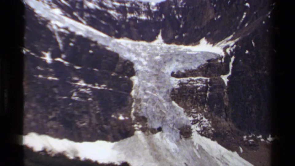 Close 8mm view of a mountain waterfall in British Columbia, with rocky cliffs, snow patches, and dark surrounding slopes.