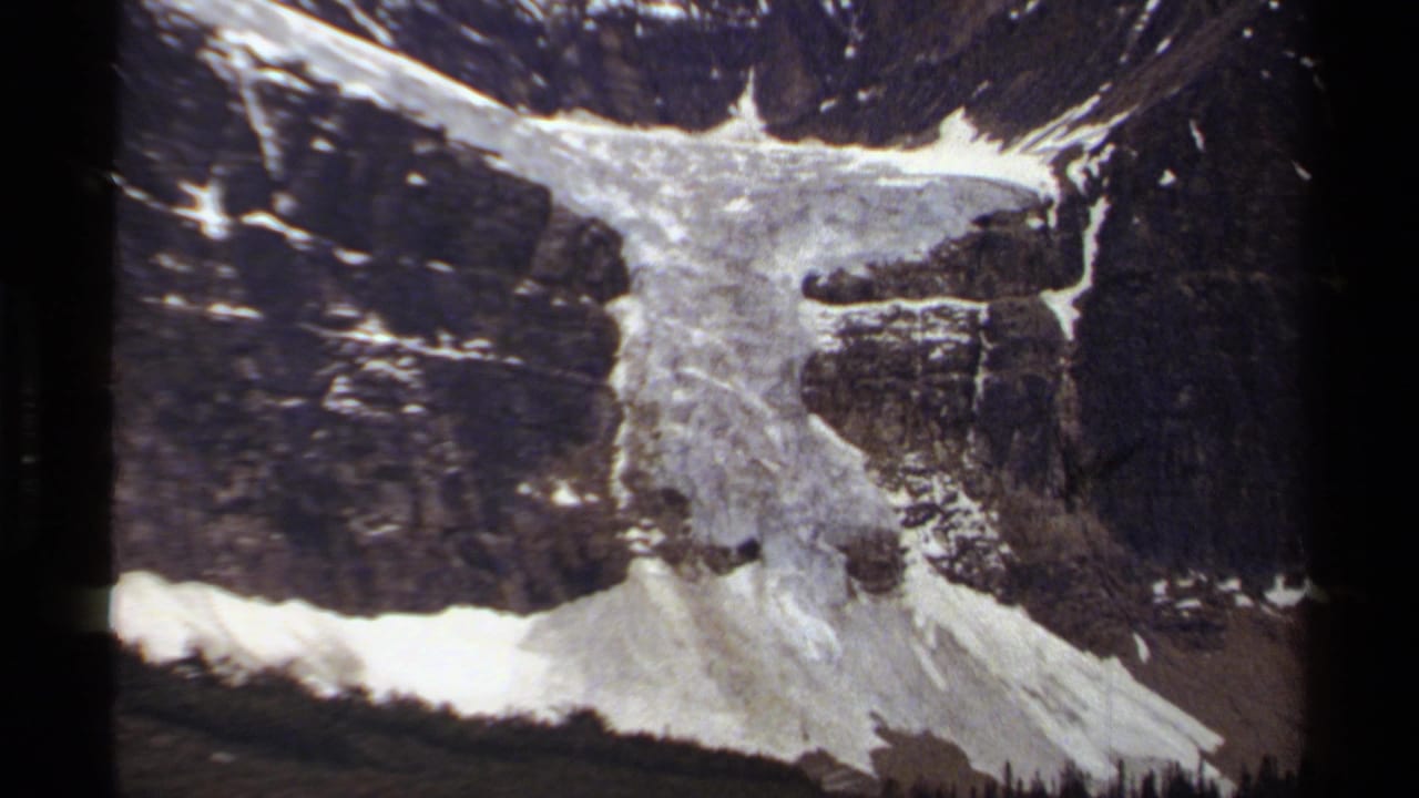 1979 8mm view of a British Columbia alpine slope with snowfields, dark cliffs, scree, and distant trees.