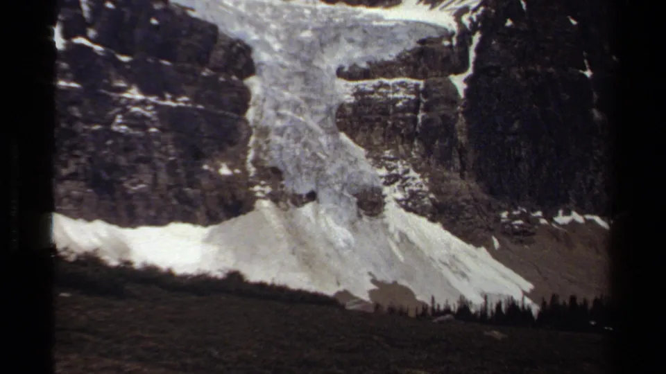 1979 home-movie frame of a snow-covered British Columbia mountain side with cliffs, scree, and distant trees.