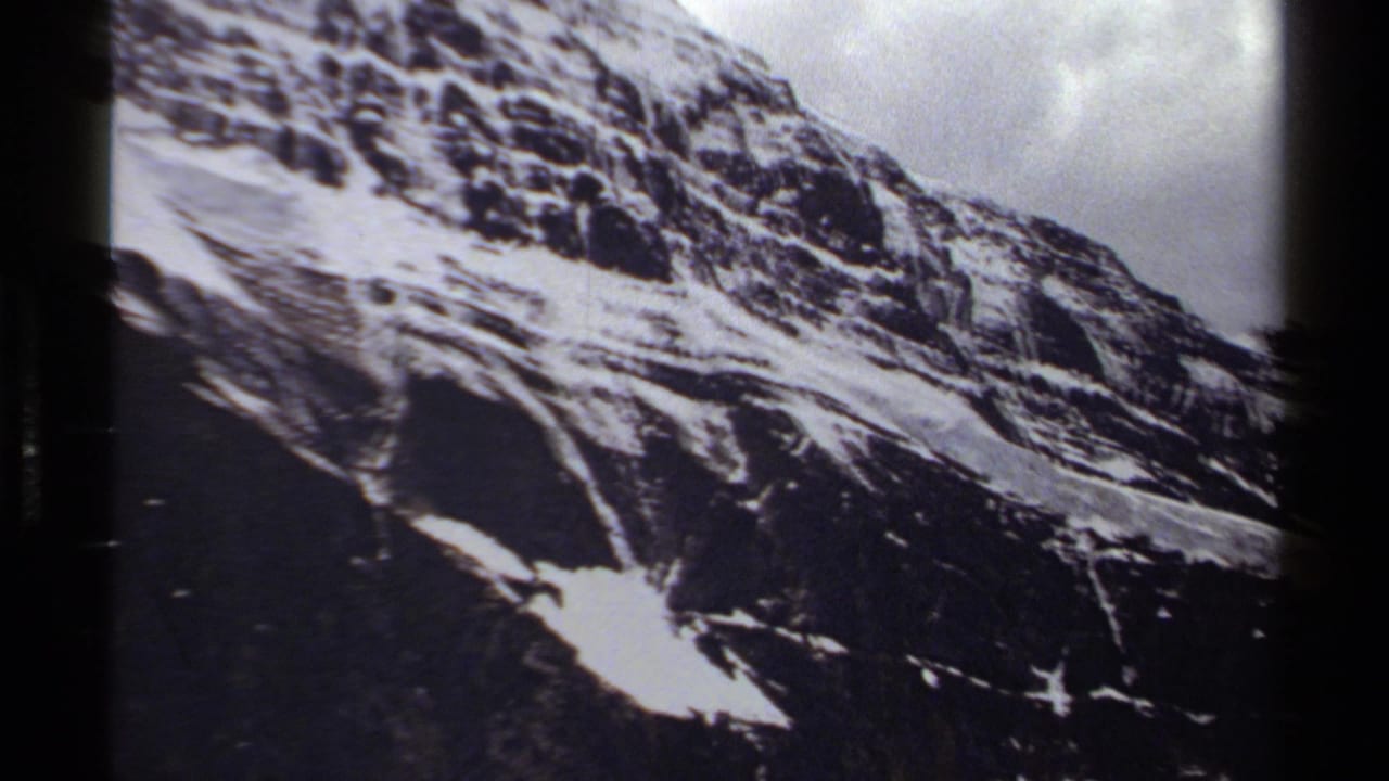 1979 British Columbia 8mm frame showing a dark rocky mountain slope with patches of snow and cloudy sky.