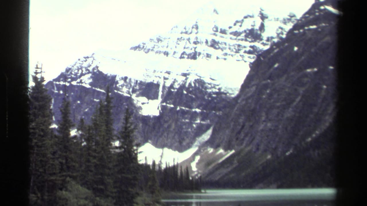 Snow-capped mountain above evergreen trees and a pale alpine lake.