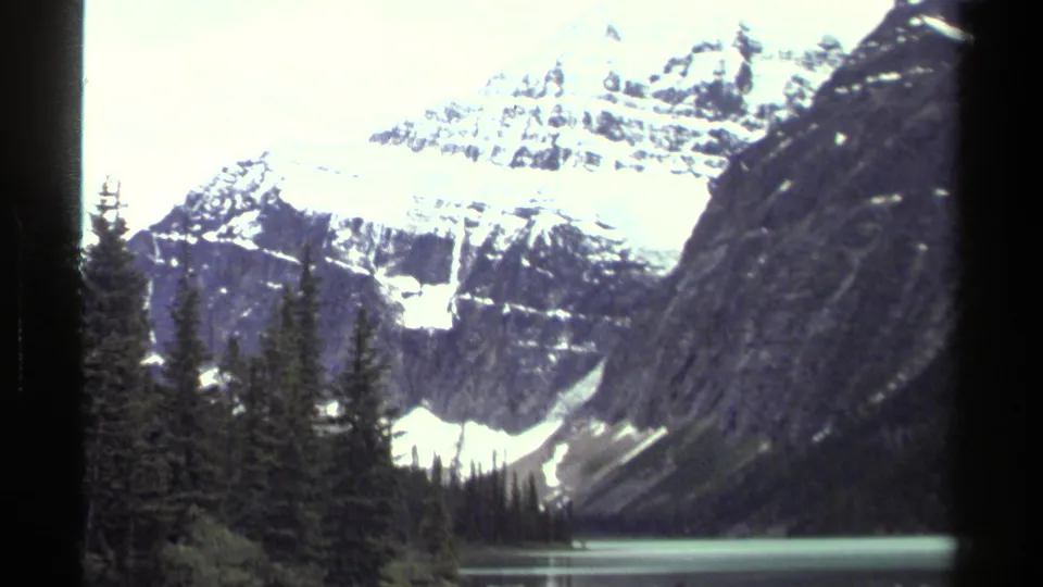 1979 8mm view of a snow-capped mountain, dark evergreens, rocky slopes, and a calm alpine lake in the Canadian travel footage.