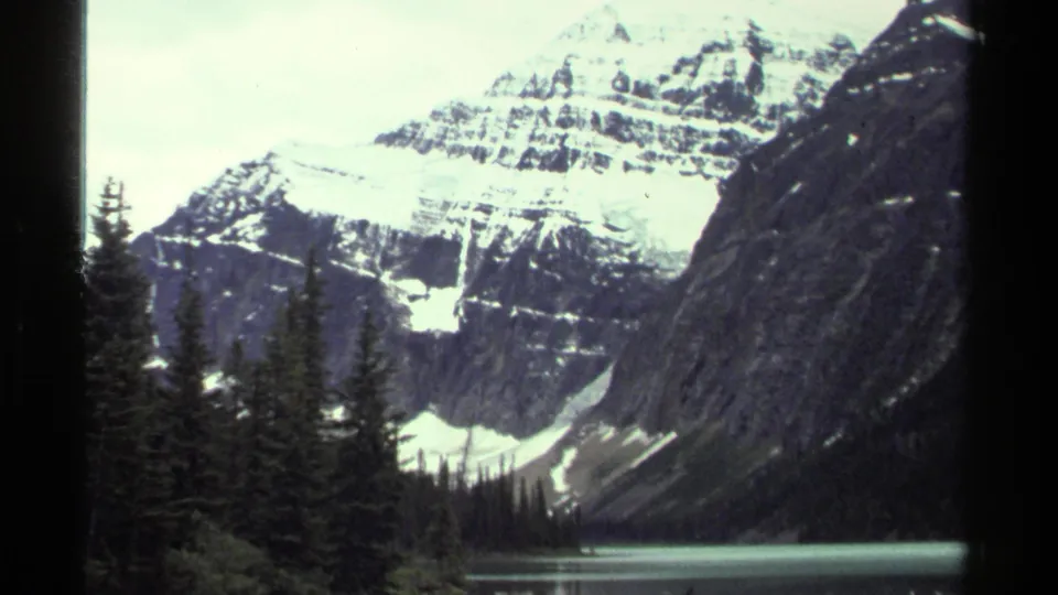 Vintage 8mm frame of a snow-covered mountain and alpine lake, with evergreen trees and steep dark slopes in the foreground.
