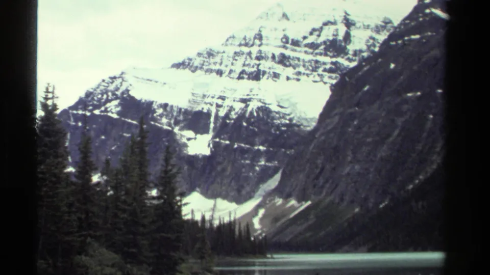 1979 8mm travel frame showing snow-covered peaks above evergreens and a narrow strip of pale alpine water.