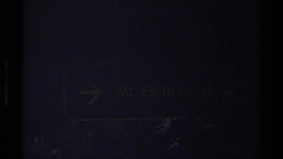 Underexposed 8mm frame of a brown and yellow directional sign reading Mt. Edith Cavell against a dark forested background.
