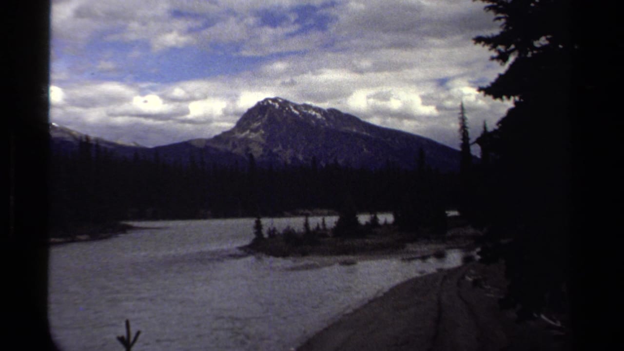 1979 8mm view of a lake and evergreen shoreline with a snow-streaked mountain peak under a cloudy sky.