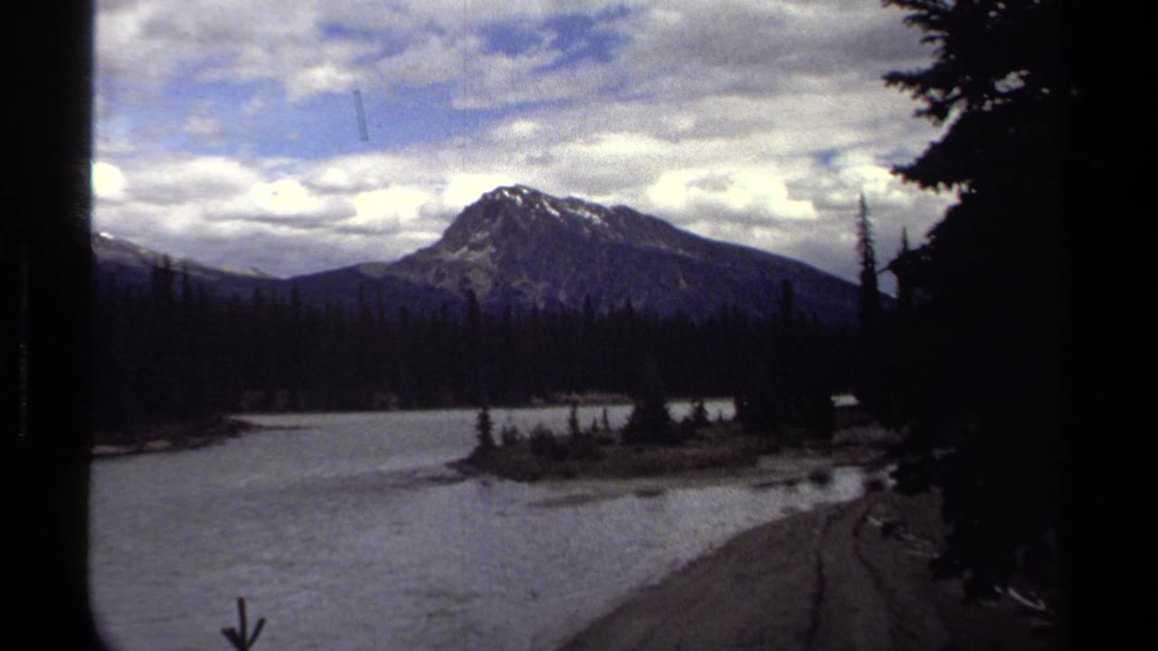 Canadian Rockies travel frame showing a lake, dark pines, and a distant snow-streaked mountain beneath broken clouds.