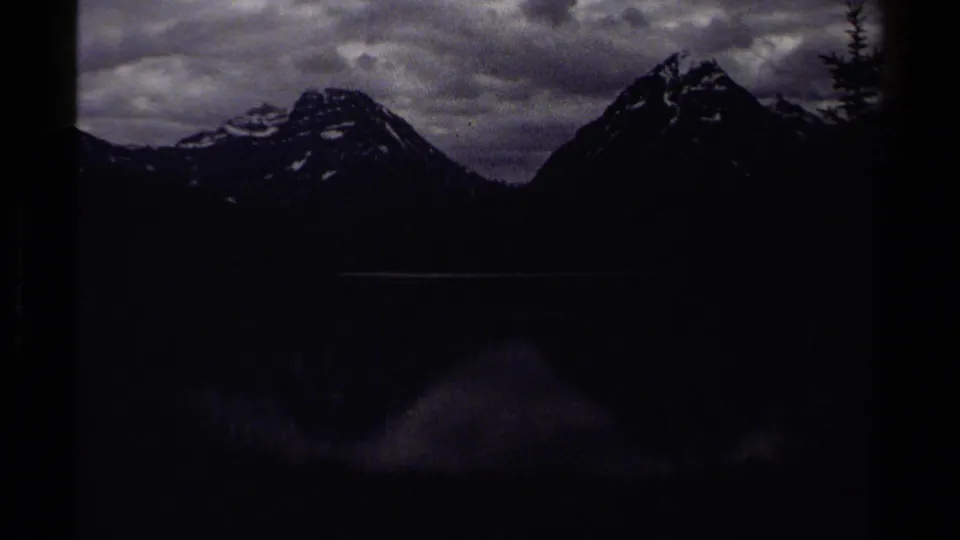 1979 British Columbia 8mm frame showing dark mountain silhouettes above a calm lake, with cloudy sky and heavy film-edge shadowing.