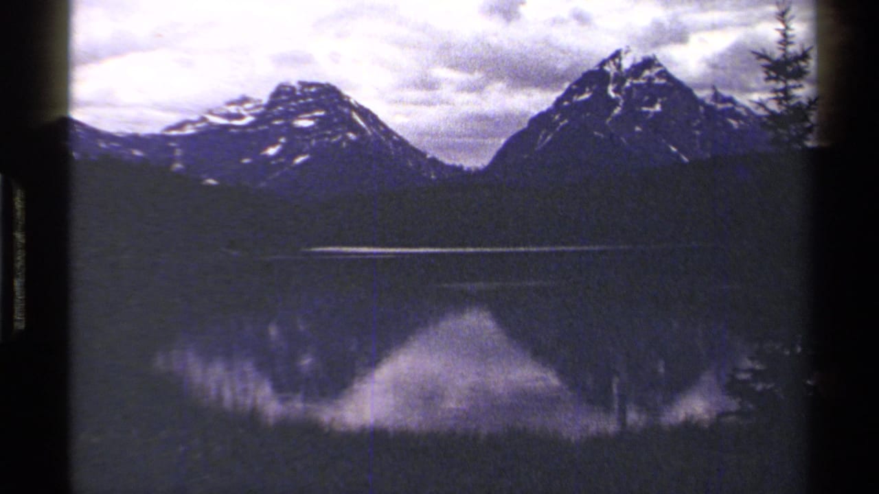 Snow-streaked mountains reflected in a still lake with cloudy sky above.