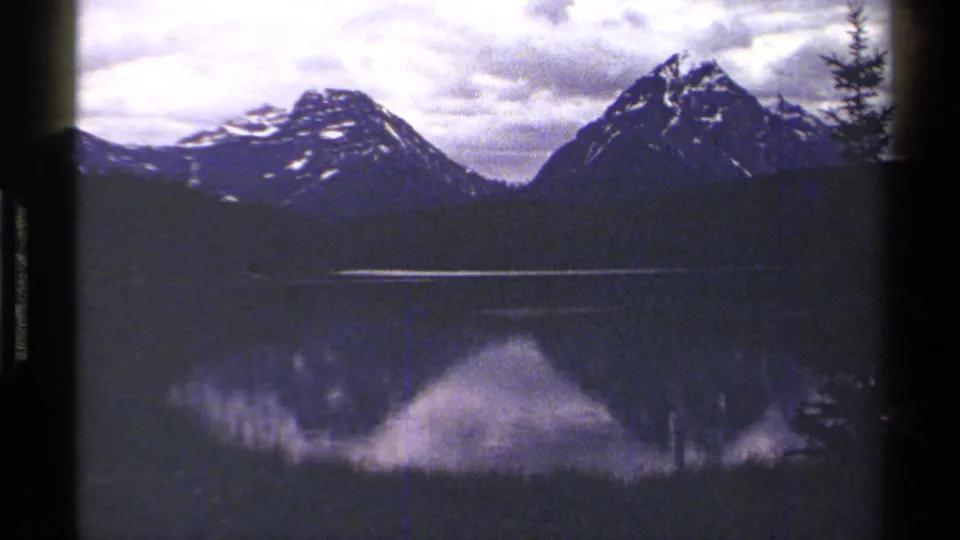 1979 British Columbia 8mm view of snow-marked peaks and forested shoreline reflected in a calm lake beneath a cloudy sky.