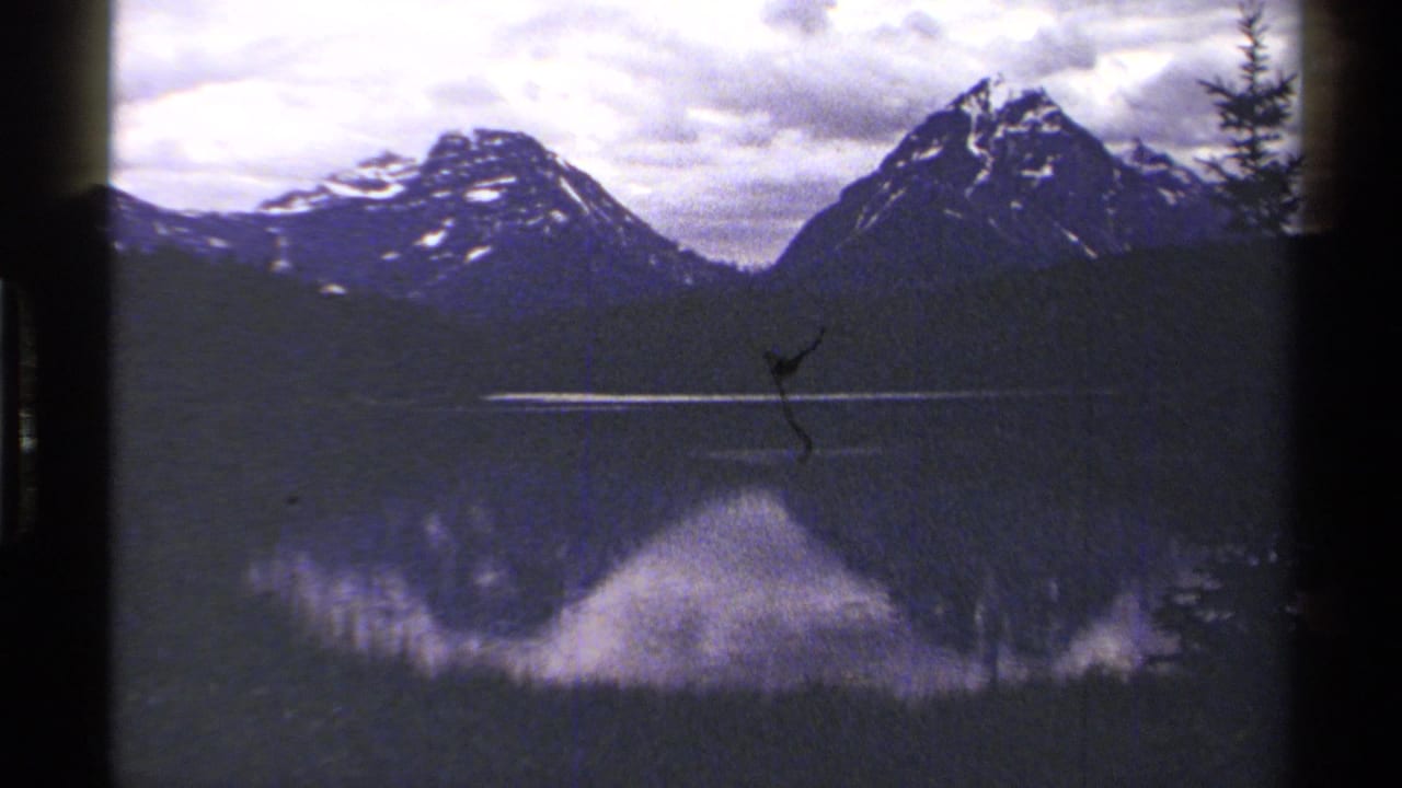 1979 British Columbia 8mm frame showing a mountain lake reflection, cloudy sky, dark treeline, and a small film scratch near the center.