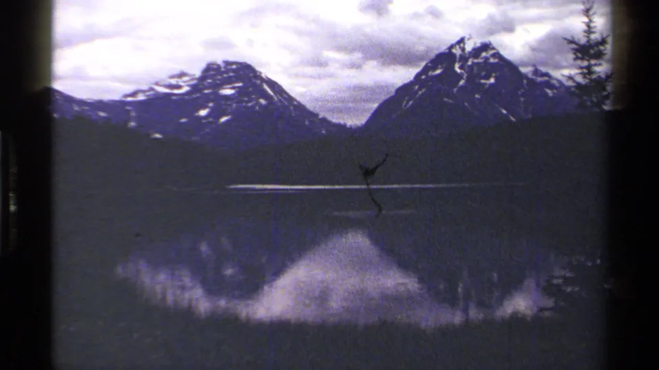 1979 British Columbia 8mm frame showing a mountain lake reflection, cloudy sky, dark treeline, and a small film scratch near the center.