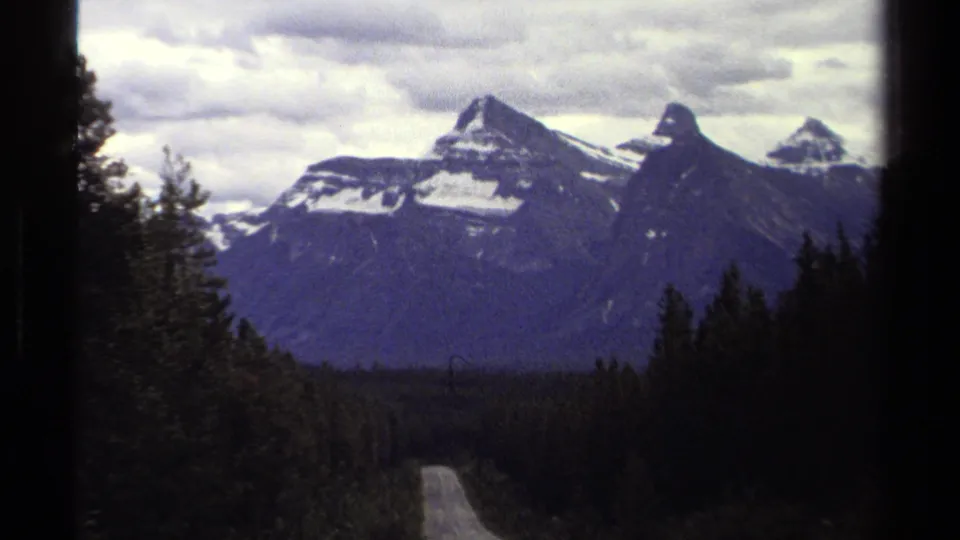 British Columbia home-movie frame with a narrow road, dense trees, and snow-dusted mountain peaks in soft 8mm color.
