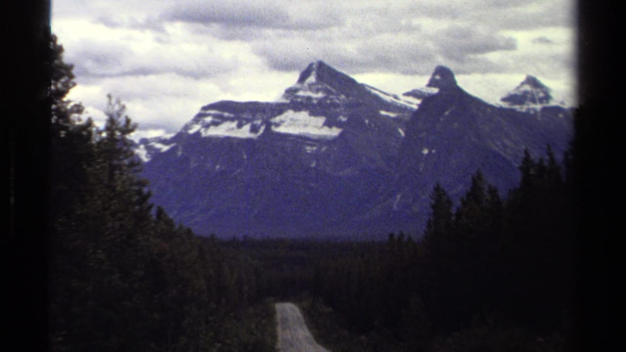 8mm travel frame from British Columbia showing a curving road through dark conifers toward snow-marked mountains.