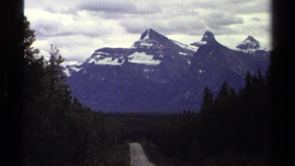 8mm travel frame from British Columbia showing a curving road through dark conifers toward snow-marked mountains.