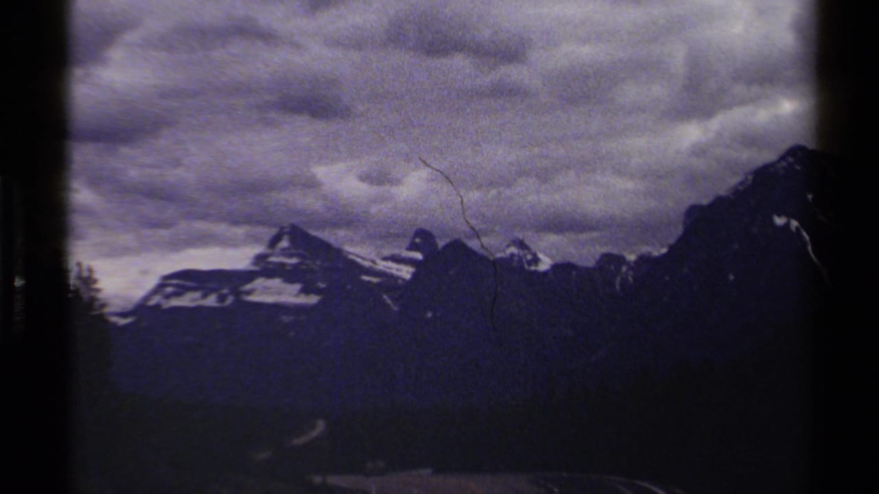 British Columbia 8mm frame with a darker mountain ridge, clouded sky, forested valley, and a small pale road in the distance.