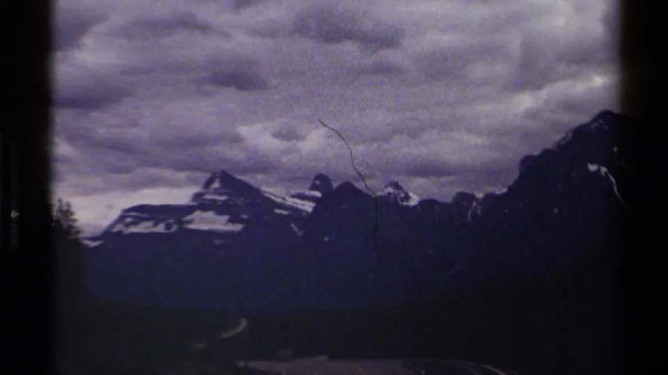 British Columbia 8mm frame with a darker mountain ridge, clouded sky, forested valley, and a small pale road in the distance.
