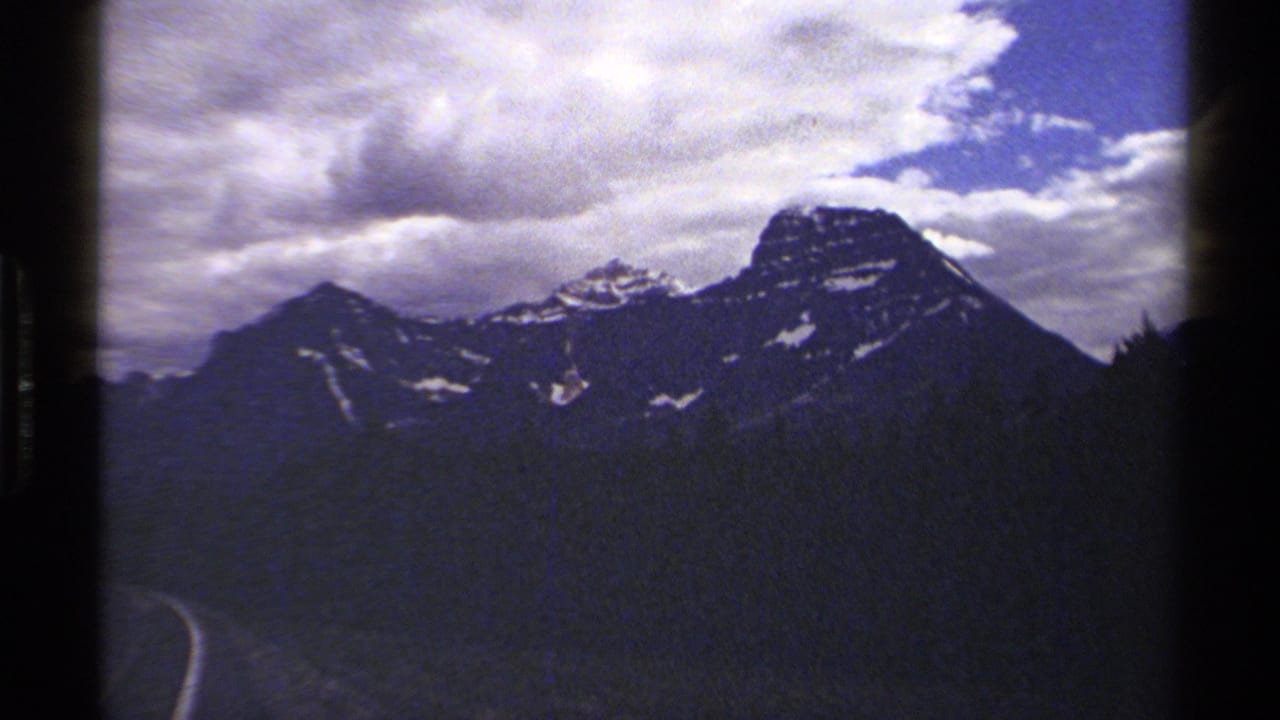 A 1979 8mm view of British Columbia mountains with dark forested slopes and a road curve entering the lower edge of the frame.