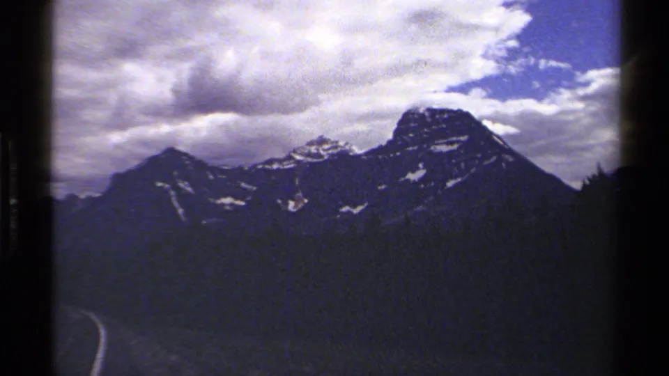 A 1979 8mm view of British Columbia mountains with dark forested slopes and a road curve entering the lower edge of the frame.