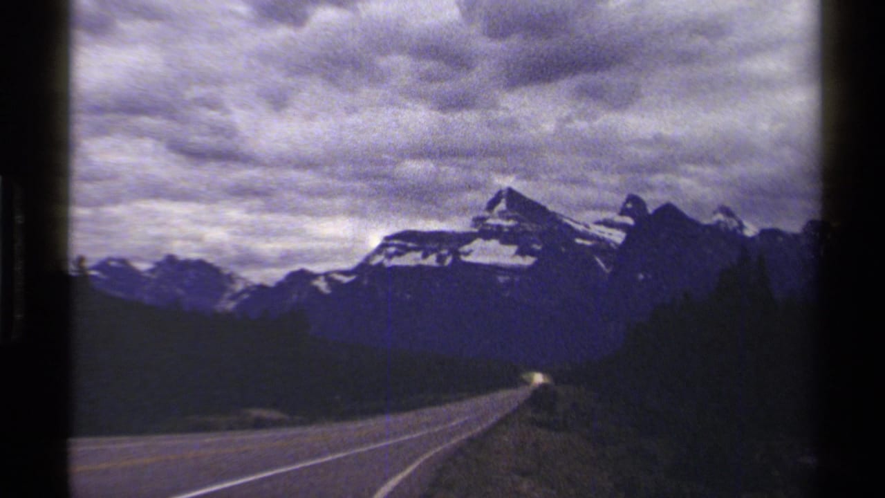 A paved highway runs toward distant British Columbia mountains in 1979 8mm travel footage, with clouds and forested edges visible.