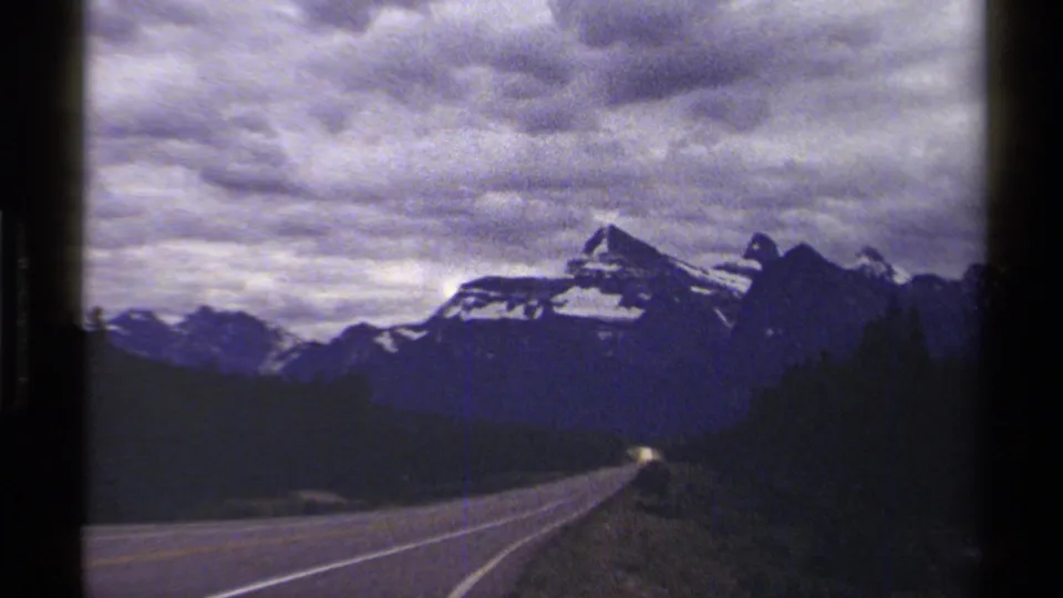 A paved highway runs toward distant British Columbia mountains in 1979 8mm travel footage, with clouds and forested edges visible.