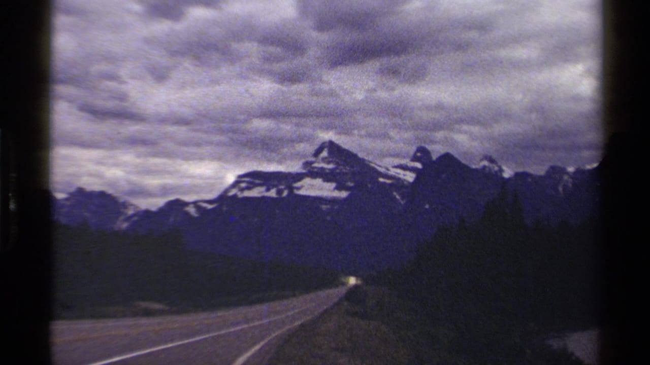 1979 8mm British Columbia road view toward snowy mountains, with forested roadside and heavy cloud cover.