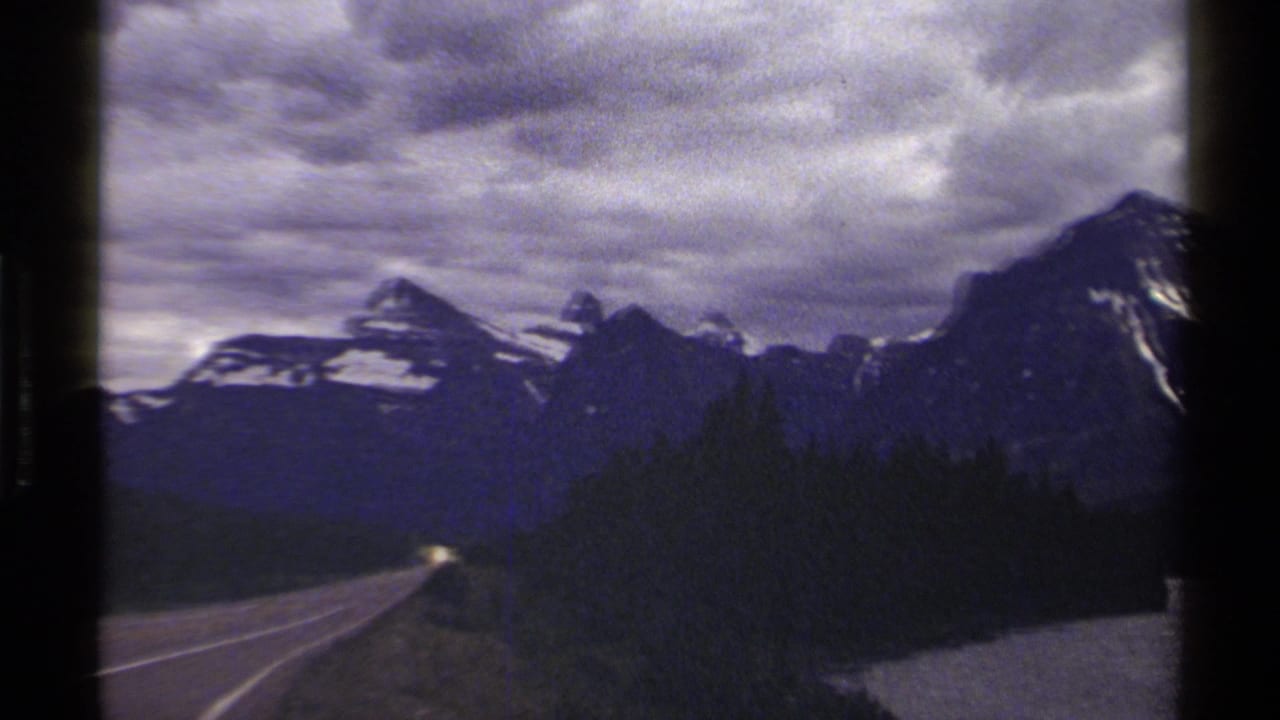Mountain highway view with dark trees and snowy peaks in the distance.