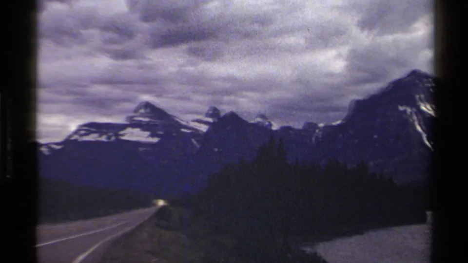 Moving 8mm view along a British Columbia highway, with evergreen forest and snow-streaked mountain peaks ahead.