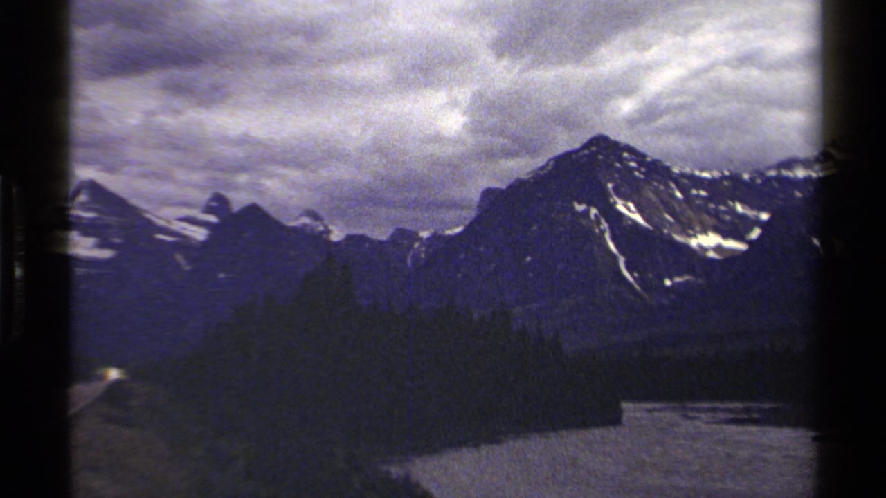British Columbia mountain scenery on 8mm film, showing forest, a partial highway edge, and a river below the peaks.