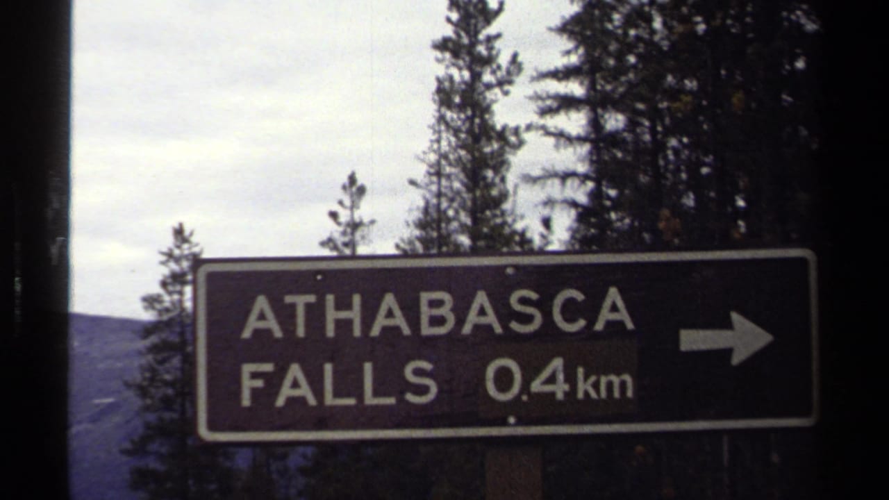 1979 8mm travel frame showing an Athabasca Falls 0.4 km direction sign with arrow, trees, and cloudy sky behind it.