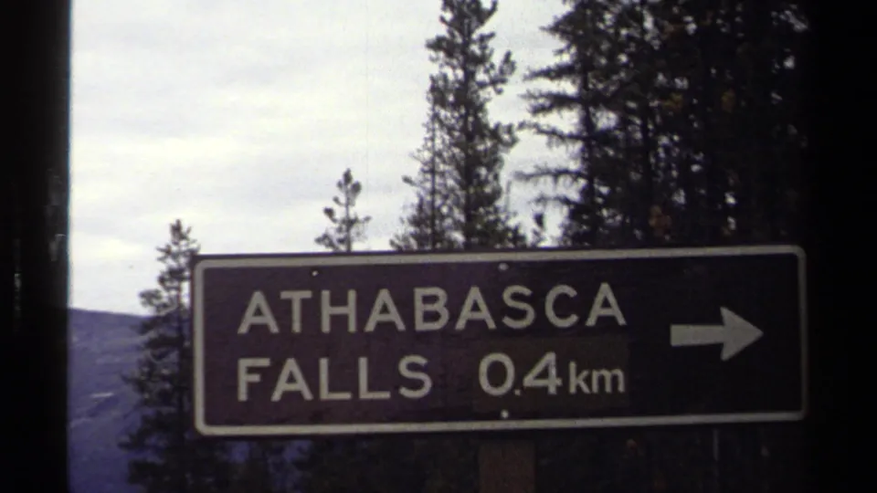 1979 8mm travel frame showing an Athabasca Falls 0.4 km direction sign with arrow, trees, and cloudy sky behind it.
