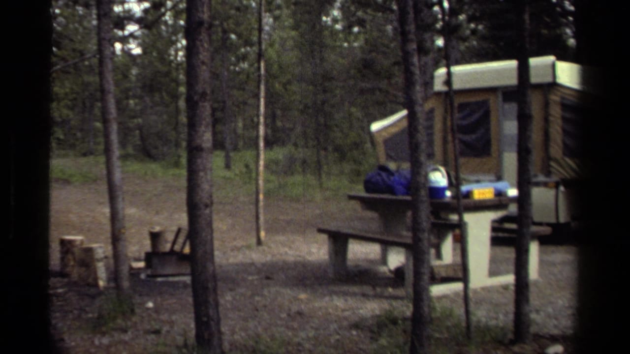 Wooded Canadian campsite with a tan pop-up camper, picnic table, and camping gear in a 1979 home-movie frame.