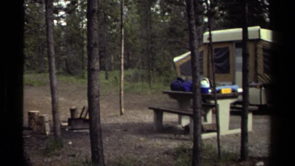 Wooded Canadian campsite with a tan pop-up camper, picnic table, and camping gear in a 1979 home-movie frame.