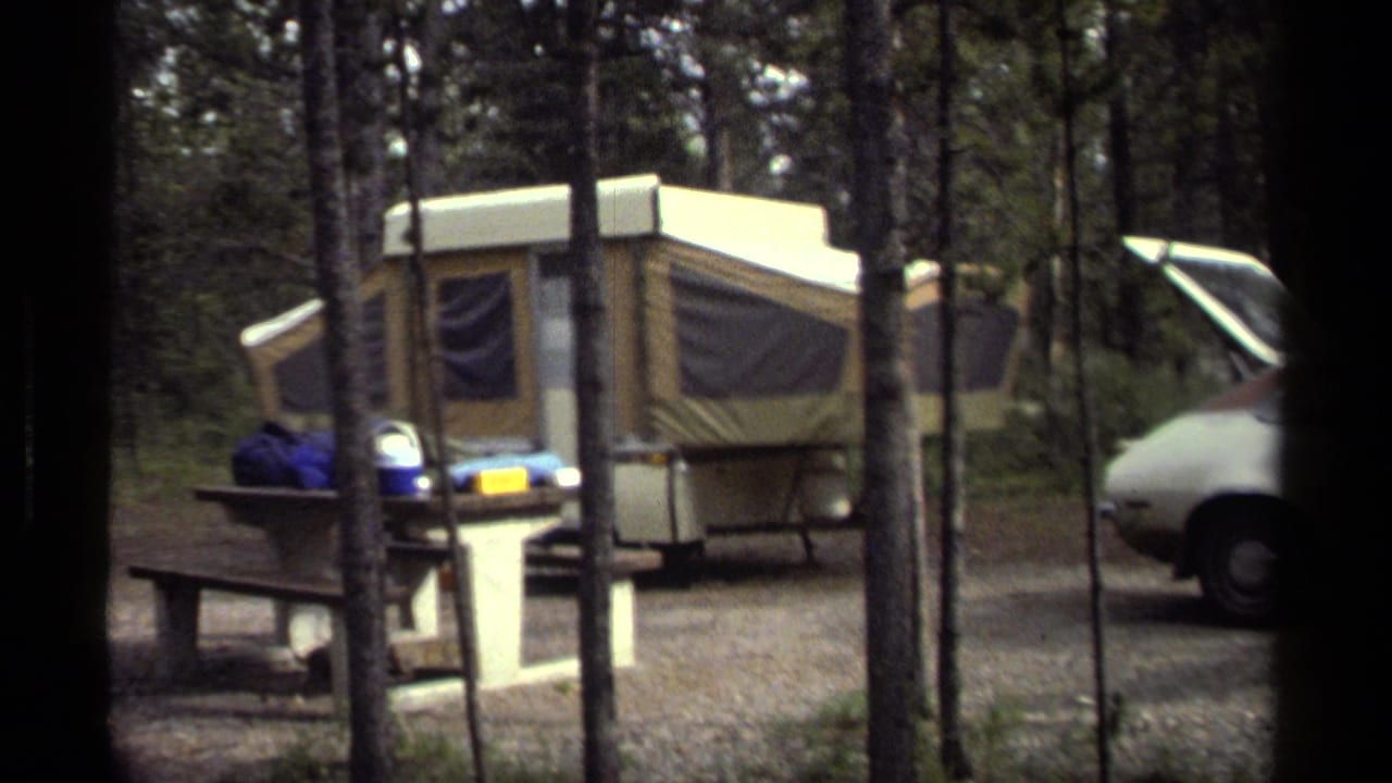 1979 campground view showing a tan pop-up camper, picnic table, trees, and part of a parked older car.