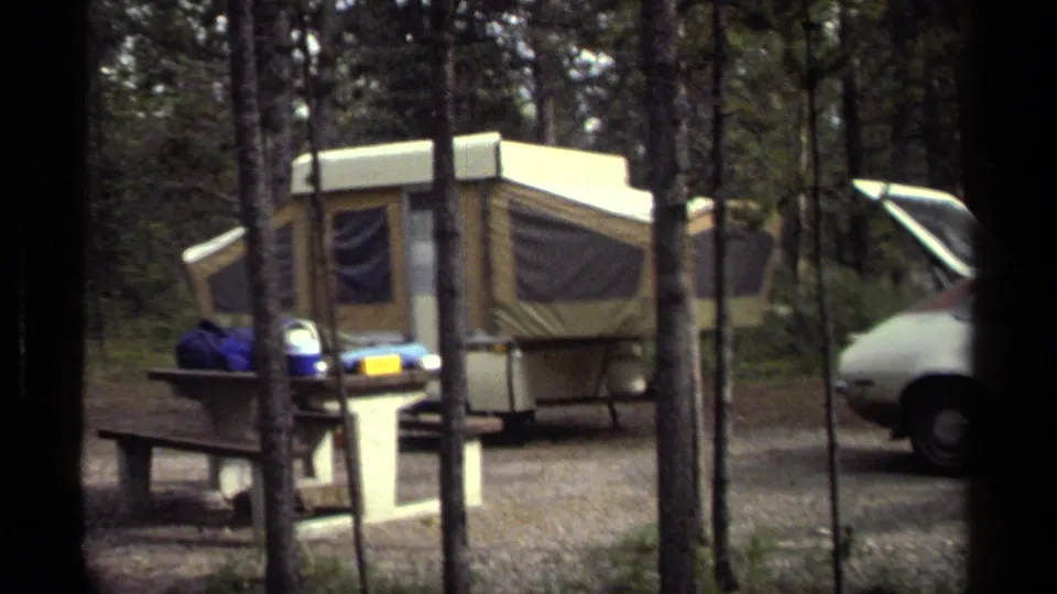 1979 campground view showing a tan pop-up camper, picnic table, trees, and part of a parked older car.