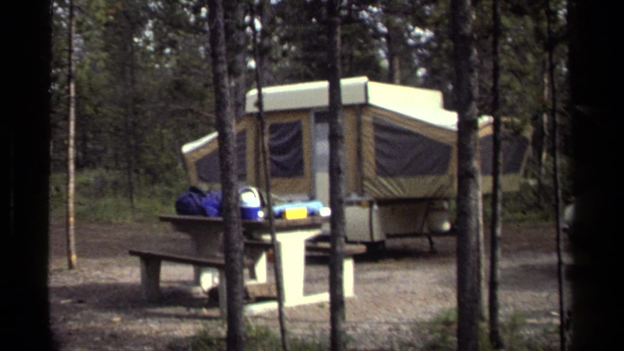 1979 British Columbia 8mm frame showing a tan pop-up camper, picnic table, and camping gear among tall trees.