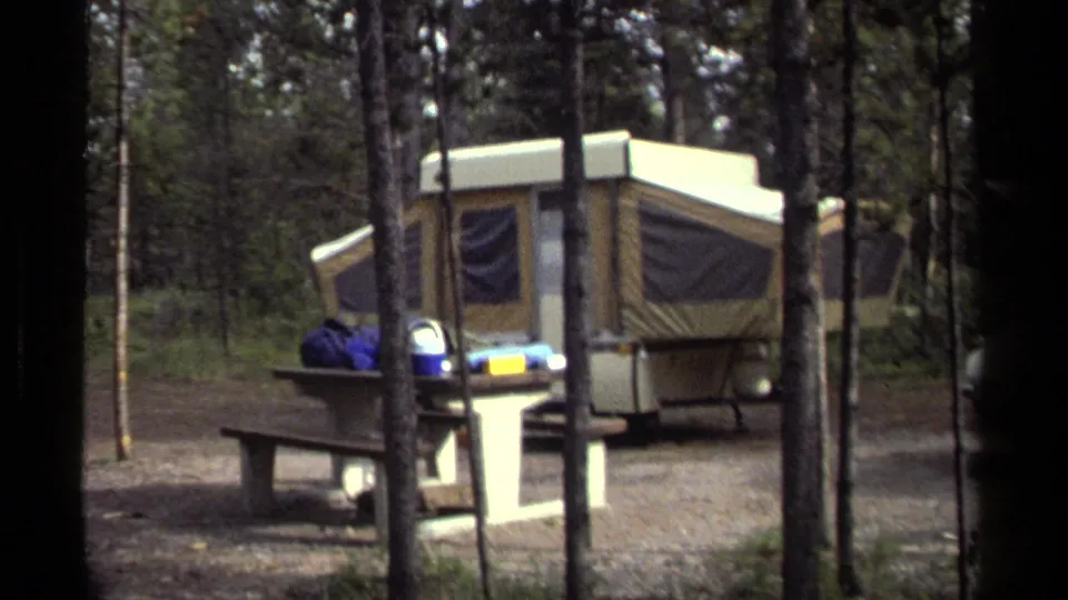 1979 British Columbia 8mm frame showing a tan pop-up camper, picnic table, and camping gear among tall trees.