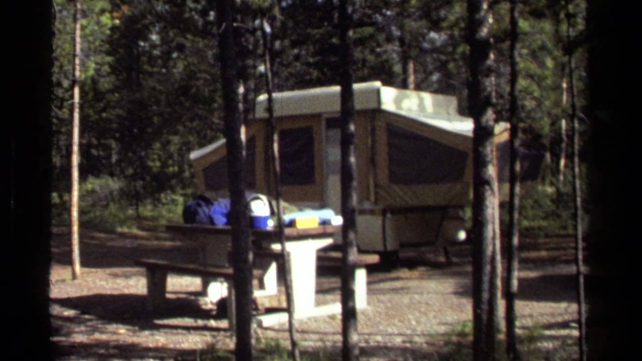 1979 8mm view of a forest campground in British Columbia with a pop-up camper and picnic table seen past trees.