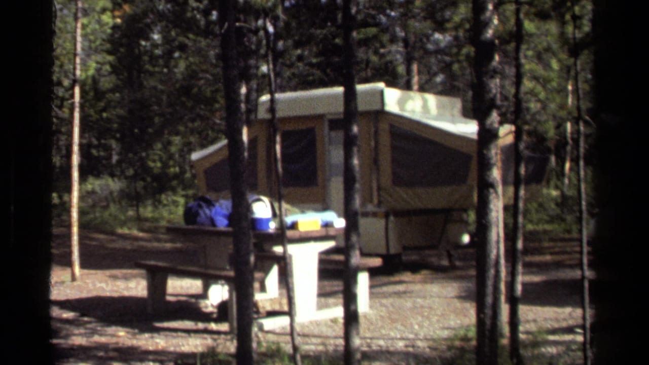 1979 British Columbia home-movie frame showing a pop-up camper and picnic table set among tall conifers at a campsite.