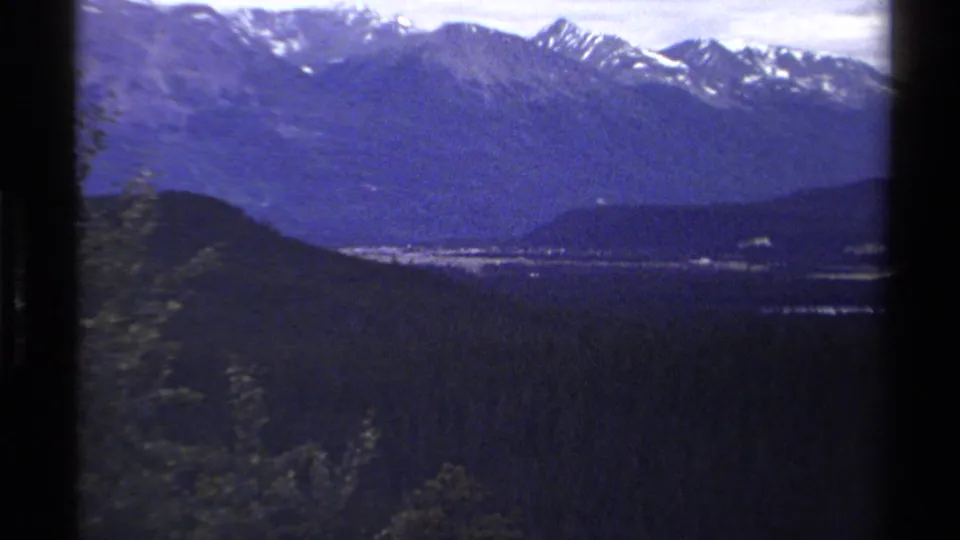 British Columbia mountain view with snow-capped peaks, forested slopes, and a distant valley or waterway in 1979 8mm footage.