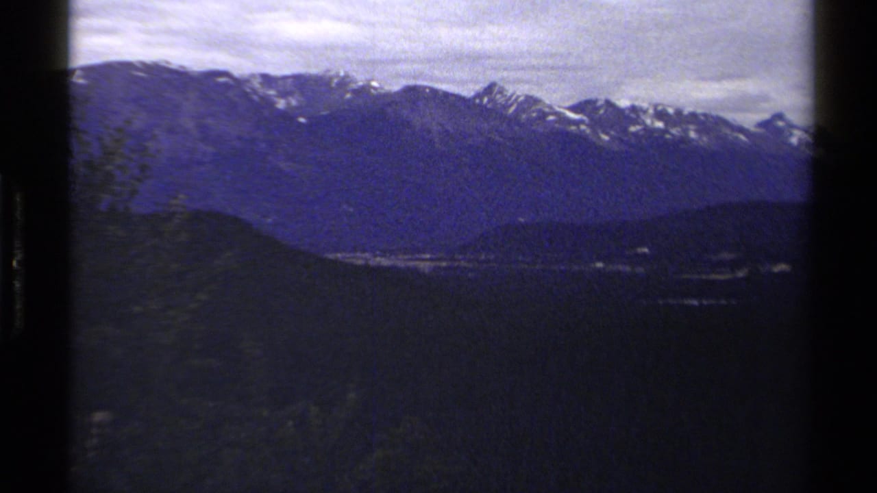 Wide 1979 British Columbia home-movie view of snowy mountain peaks over dark forested terrain under a cloudy sky.
