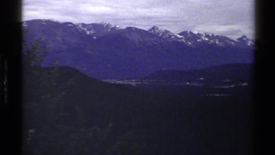 Wide 1979 British Columbia home-movie view of snowy mountain peaks over dark forested terrain under a cloudy sky.