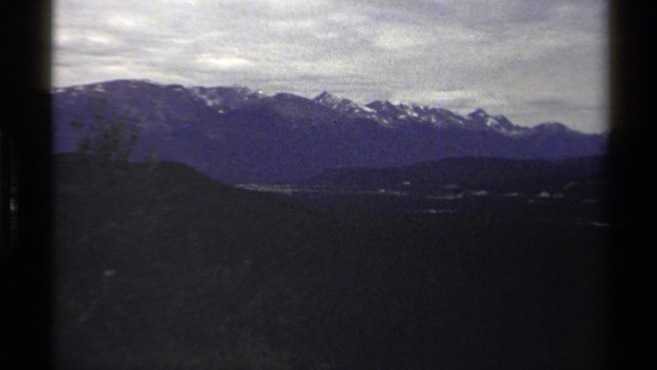 1979 British Columbia 8mm view of blue-toned mountain ridges with snow patches under a cloudy sky and dark foreground slopes.