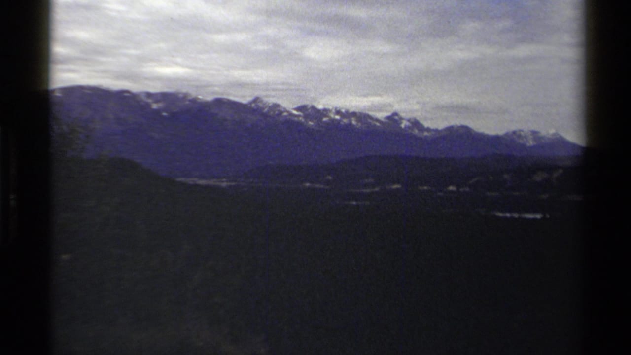 Archival 8mm frame from British Columbia showing layered mountain ridges, snow patches, and a shadowed valley under heavy cloud.