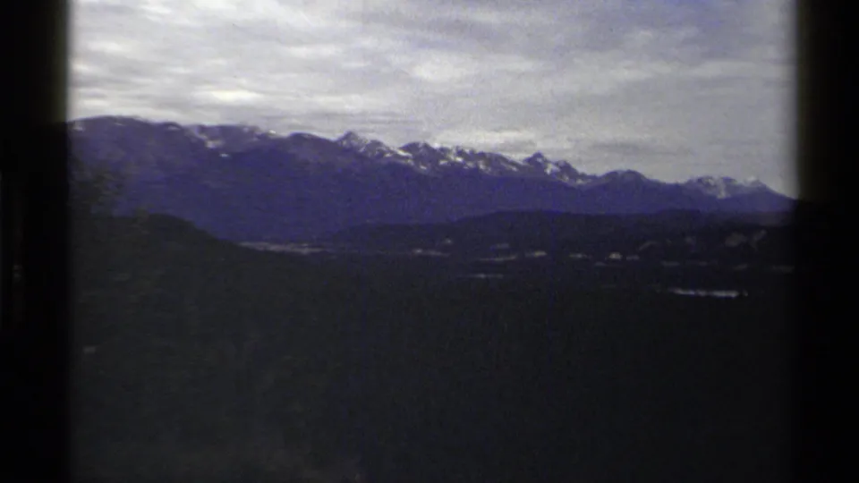 Archival 8mm frame from British Columbia showing layered mountain ridges, snow patches, and a shadowed valley under heavy cloud.
