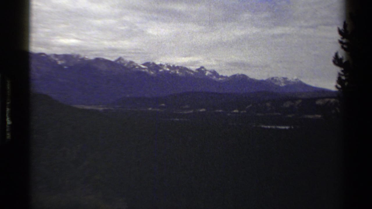 1979 8mm travel frame with British Columbia mountain peaks, cloudy sky, dark foreground terrain, and a tree silhouette at the right edge.
